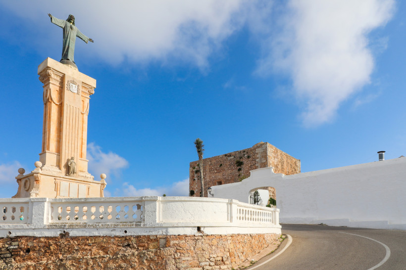 Statue von Christus mit ausgebreiteten Armen auf einem hohen Sockel am Monte Toro auf Menorca, daneben eine alte Festungsmauer unter blauem Himmel.
