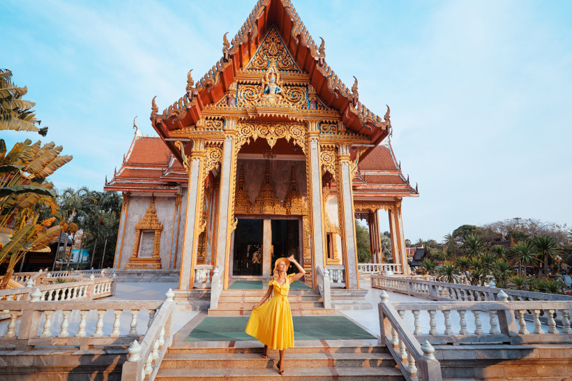 Thailand - Phuket Eine Frau in einem leuchtend gelben Kleid steht auf der Treppe vor einem prachtvollen thailändischen Tempel mit kunstvoll vergoldeten Ornamenten und traditioneller Architektur