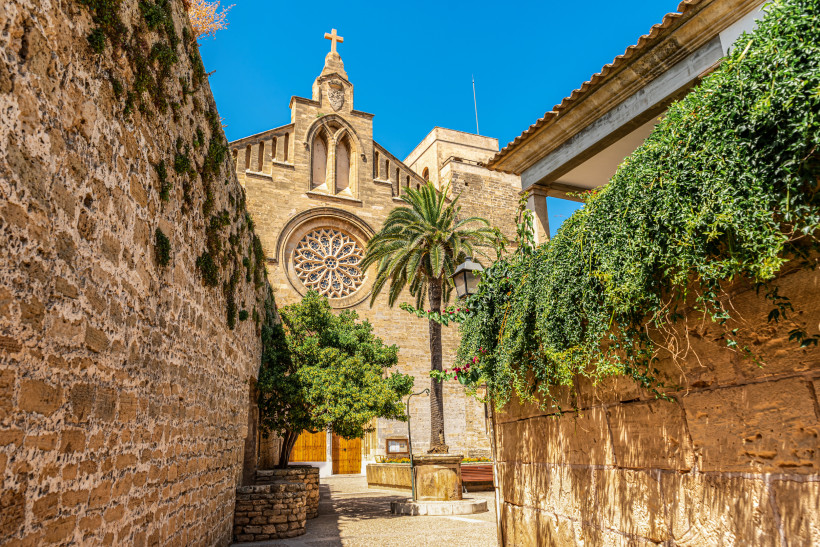 Alcúdia, Mallorca Historische Kirche mit Rosettenfenster und Kreuz in Alcúdia auf Mallorca, umgeben von Steinmauern, Palmen und blauem Himmel