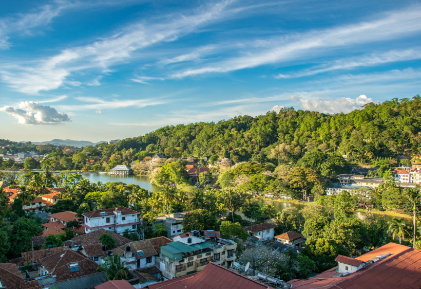 Panoramaaufnahme der Stadt Kandy in Sri Lanka, mit einem See im Zentrum, umgeben von tropisch grünen Hügeln. Im Vordergrund Dächer der Stadt mit roten Ziegeln, im Hintergrund dichter Wald und einzelne Gebäude. Der Himmel ist blau mit leichten Wolken, die 