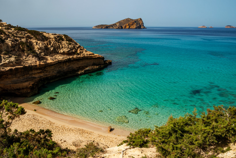 Bucht bei Cala Comte mit Sandstrand, Felsklippe und türkisfarbenem Meer, im Hintergrund eine kleine Insel