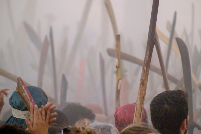 Mallorca Nahaufnahme von Menschen mit erhobenen Holzstöcken in einer dichten Rauchwolke, aufgenommen während des Festes 'Moros i Cristians' in Sóller. Viele Teilnehmer tragen Kopfbedeckungen oder bunte Kleidung. Die Szene wirkt intensiv und emotional – symbolträch