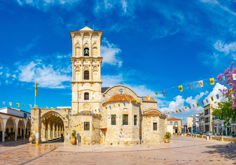 Kirche des Heiligen Lazarus in Larnaca: helles Sandsteinbauwerk mit markantem Glockenturm, Arkadengang und Wimpelketten auf dem Vorplatz.