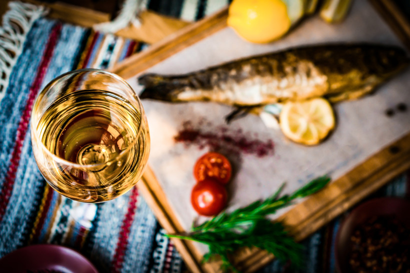 Ägypten - Sharm el Sheikh Ein gedeckter Tisch mit einem Glas Weißwein im Vordergrund und einem gegrillten ganzen Fisch im Hintergrund, serviert auf einem Holzbrett mit Zitronenscheiben, Tomaten, Rosmarin und Gemüse. Der Tisch ist mit einem bunten, gewebten Tuch bedeckt, die Atmosp
