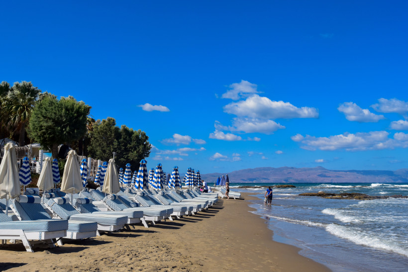 Familie am Strand von Platanias zwischen Liegen und Sonnenschirmen Strand von Platanias mit Reihen von Liegen und blau-weiß gestreiften Sonnenschirmen, Wellen am Ufer und Bergen am Horizont