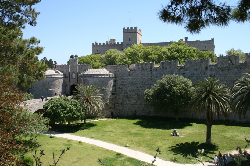 Das Bild zeigt den Großmeisterpalast der Johanniter in Rhodos-Stadt auf der griechischen Insel Rhodos.   Im Vordergrund liegt ein weitläufiger Garten mit Palmen, Bäumen, Rasenflächen und einem geschwungenen Weg.   Dahinter ragen hohe Burgmauern aus grauem