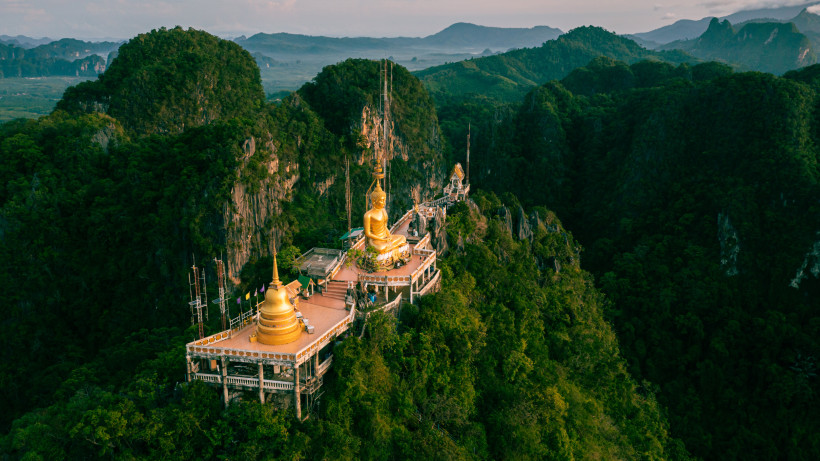 Tiger Cave Temple in Krabi, Thailand: Luftaufnahme des Tempels mit goldener Buddha-Statue auf Berggipfel inmitten tropischer Landschaft.