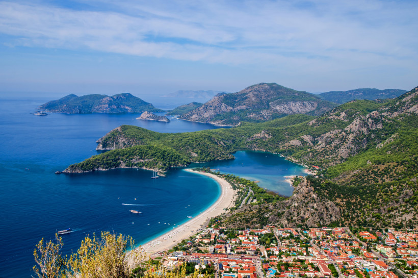 Luftaufnahme der Küstenlandschaft von Ölüdeniz an der Türkischen Riviera mit einer geschwungenen Sandbank, türkisblauem Meer, grünen Bergen und vorgelagerten Inseln.