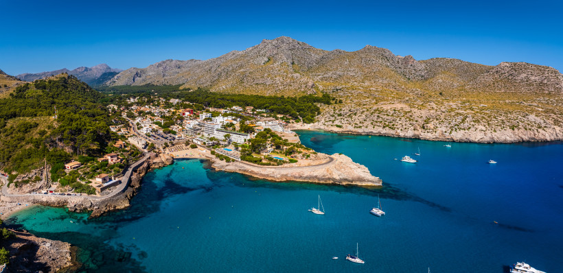 Luftaufnahme von Cala Sant Vicenç mit Küstenort, kleinen Sandbuchten, türkisblauem Meer und Booten vor einer Bergkulisse
