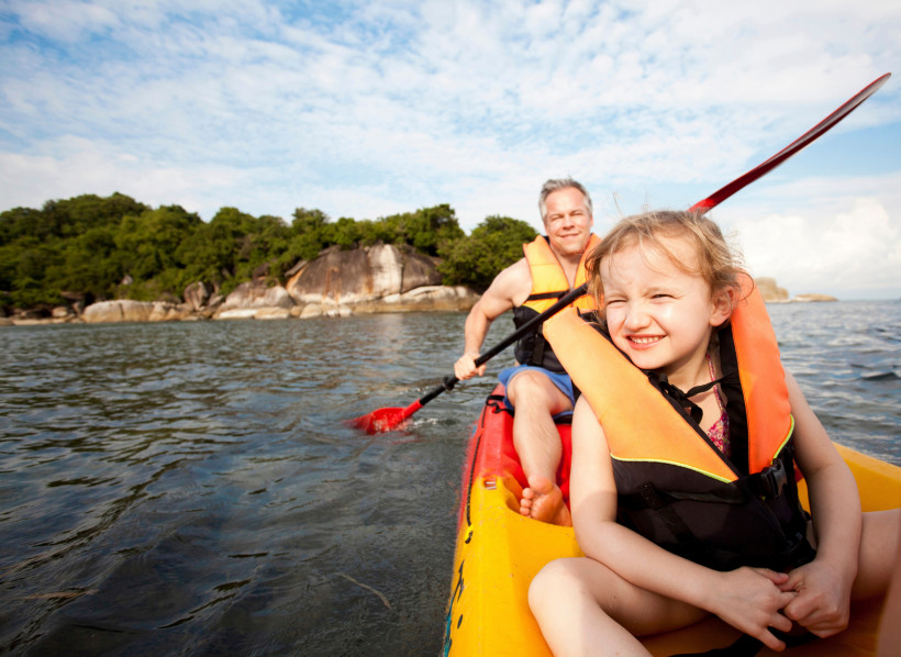 Thailand - Koh Samui Das Bild zeigt einen Mann und ein kleines Mädchen, die zusammen in einem farbenfrohen Kajak auf dem Wasser paddeln. Beide tragen orangefarbene Schwimmwesten, der Mann sitzt hinten mit einem Paddel in der Hand, während das Mädchen vorne sitzt und fröhlich
