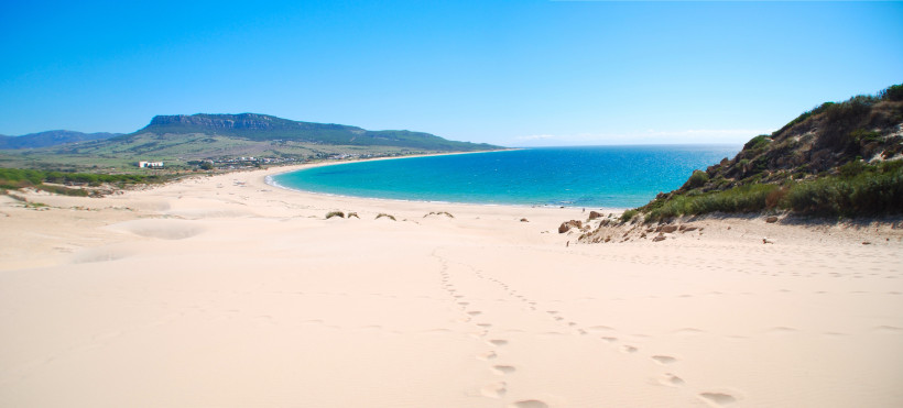 Weißer Sandstrand Playa de Bolonia an der südspanischen Atlantikküste. Der Blick fällt über Dünen auf das türkisfarbene Meer und eine langgezogene Bucht, eingerahmt von grünen Hügeln und Felsen unter wolkenlosem Himmel.