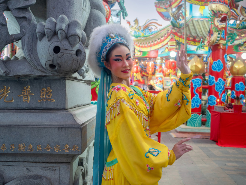 Thailand - Phuket Frau in farbenfrohem, gelbem traditionellen Kostüm mit Kopfschmuck. Sie posiert elegant vor einem reich geschmückten chinesischen Tempel mit roten und blauen Säulen.