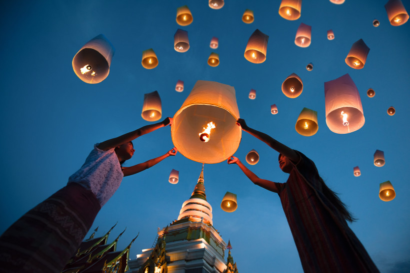 Thailand Pattaya Zwei Personen lassen gemeinsam eine leuchtende Himmelslaterne in den Abendhimmel steigen. Rundherum steigen viele weitere Laternen auf, im Hintergrund ein thailändischer Tempel mit goldener Spitze.