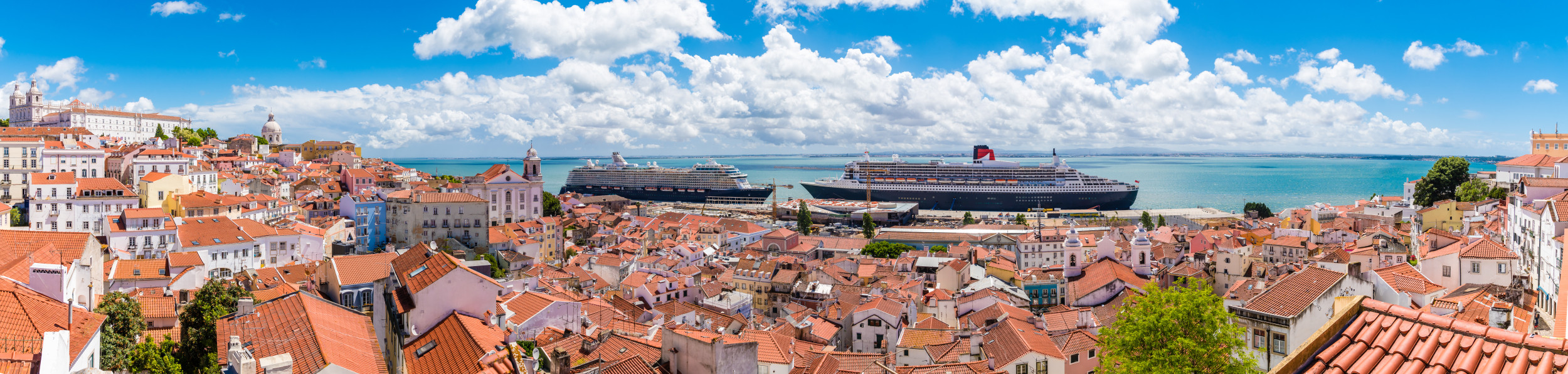 Blick über die Altstadt von Lissabon mit roten Ziegeldächern und zwei Kreuzfahrtschiffen im Hafen am Tejo