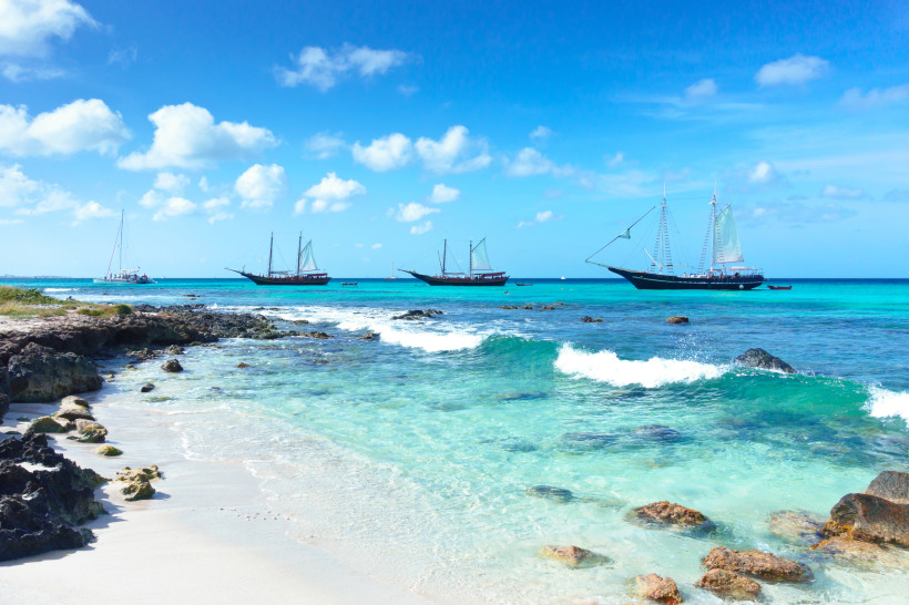 Türkisblaues Meer mit mehreren großen Segelschiffen, die vor der Küste von Aruba liegen. Im Vordergrund ein felsiger, weißer Sandstrand mit sanften Wellen und kristallklarem Wasser unter einem strahlend blauen Himmel.