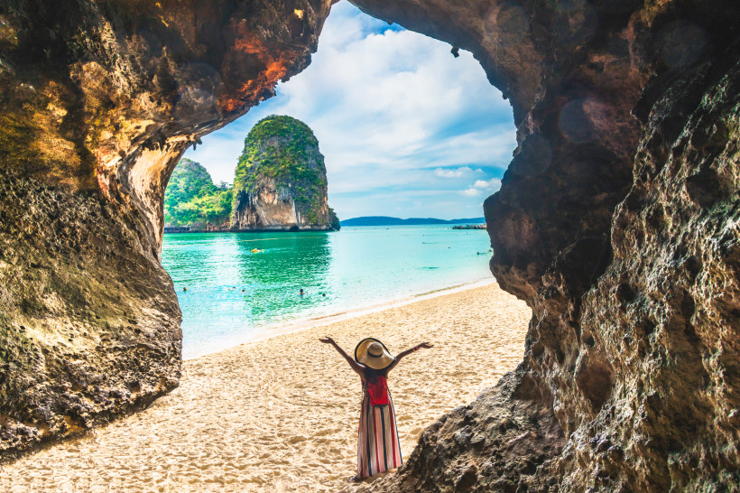 Phra Nang Cave Beach in Krabi, Thailand: Frau mit Hut blickt vom Höhleneingang auf weißen Sandstrand, türkisfarbenes Meer und Karstfelsen.