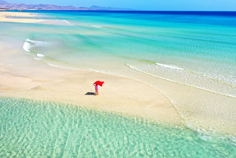 Traumstrand mit türkisfarbenem Wasser Person mit rotem Tuch steht allein auf einem flachen Sandstrand im türkisblauen, klaren Meer, mit Wellen und Bergen am Horizont unter blauem Himmel