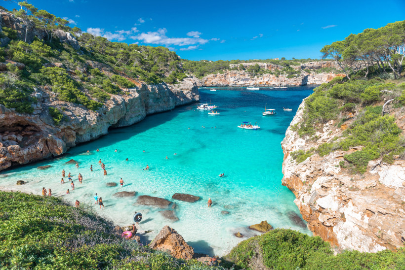 Mallorca Strand Cala des Moro auf Mallorca mit kristallklarem, türkisfarbenem Wasser, Badenden im flachen Wasser und Booten in der Bucht, umgeben von Felsen und grüner Vegetation