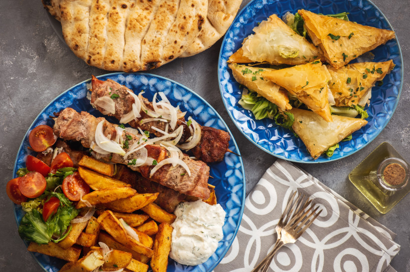 Teller mit griechischen Spezialitäten: gegrillte Fleischspieße mit Zwiebeln, Pommes frites, Tomatensalat und Tsatsiki auf einem blauen Teller. Daneben ein weiterer Teller mit goldbraun gebackenen Blätterteig-Dreiecken. Im Hintergrund Fladenbrot und eine F