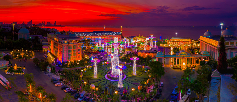 Thailand Pattaya Blick über einen hell erleuchteten Themenpark in Pattaya bei Sonnenuntergang. Der Himmel leuchtet intensiv rot und lila, während unzählige Lichter den Park in Szene setzen.