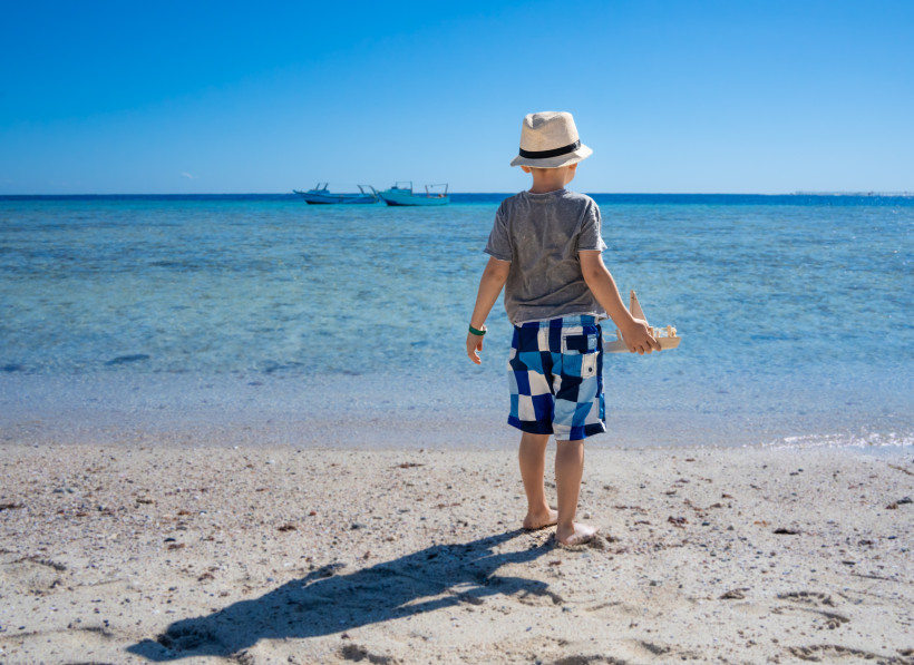 Ägypten - Marsa Alam Ein kleiner Junge steht am Strand und blickt aufs Meer. Er trägt Strohhut, T-Shirt und Badeshorts und hält ein Spielzeugboot in der Hand. Im Hintergrund sind zwei Boote auf dem Wasser zu sehen.
