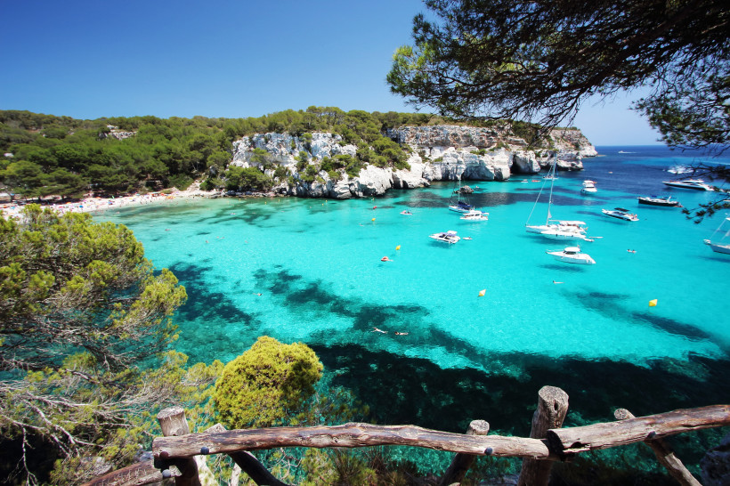 Atemberaubender Blick auf eine Bucht mit türkisfarbenem Wasser auf Menorca, Spanien. Mehrere weiße Boote und Yachten liegen vor Anker. Im Hintergrund ein heller Sandstrand, gesäumt von grünen Kiefernwäldern und hellen Kalksteinfelsen. Im Vordergrund ein r