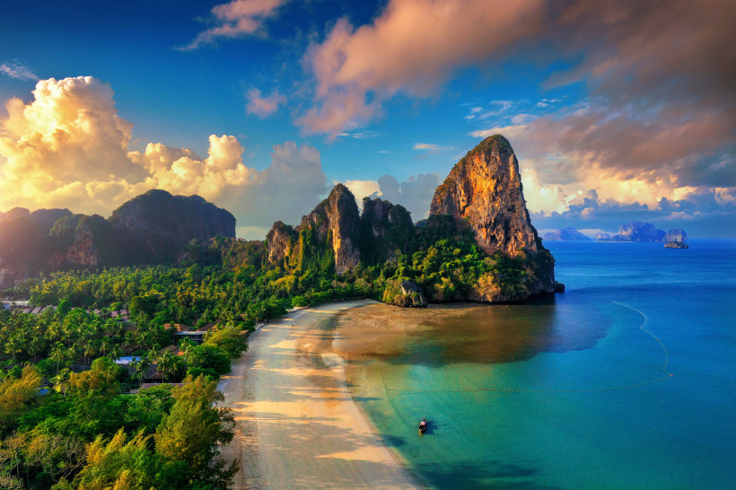 Panoramablick auf Railay Beach in Krabi bei Sonnenuntergang: goldene Wolken, türkisblaue Bucht, Longtail-Boot und steil aufragende Kalksteinfelsen an der Andamanensee.