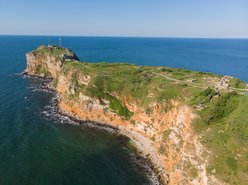 Drohnenaufnahme von Kap Kaliakra an der bulgarischen Schwarzmeerküste. Steile, rötlich gefärbte Klippen mit grüner Vegetation ragen ins Meer hinaus. Auf dem Hochplateau sind historische Ruinen und ein Leuchtturm zu erkennen.