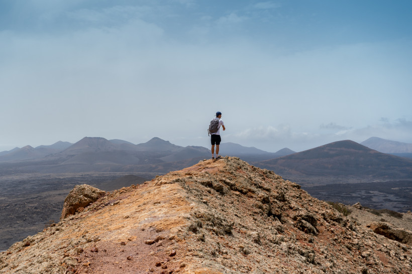 Lanzarote Wanderer mit Rucksack steht auf einem felsigen Bergrücken und blickt über eine weite Vulkanlandschaft mit sanften Hügeln unter leicht bewölktem Himmel