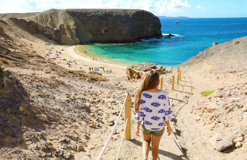Frau wandert zur Playa de Papagayo auf Lanzarote, mit goldenem Sandstrand und türkisblauem Meer in einer geschützten Felsenbucht.