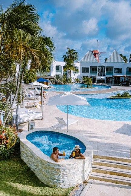 Tropische Hotelanlage auf Aruba mit weißer Fassade, Palmen und mehreren Pools. Im Vordergrund ein kleiner Whirlpool, in dem ein Paar Cocktails genießt. Im Hintergrund stehen Sonnenliegen, Sonnenschirme und weiße Gebäude mit Giebel-Dächern unter blauem Him