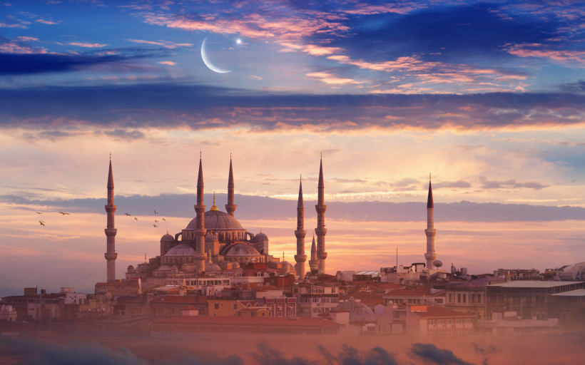 Die Silhouette der Blauen Moschee in Istanbul bei Sonnenuntergang, mit einem dramatisch gefärbten Himmel im Hintergrund. Eine Mondsichel mit einem Stern leuchtet darüber, während Vögel durch die Luft fliegen.