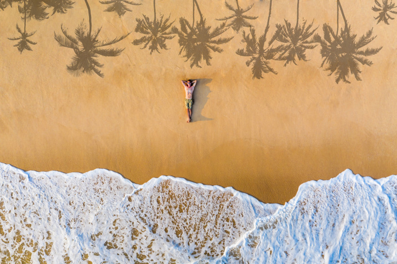 Sri Lanka Drohnenaufnahme eines Mannes, der entspannt auf dem Rücken im Sand eines goldgelben Strandes liegt. Die Brandung rollt sanft an den unteren Bildrand, während sich die Schatten von Palmen auf den Sand zeichnen. Die Szene wirkt ruhig, tropisch und friedlich