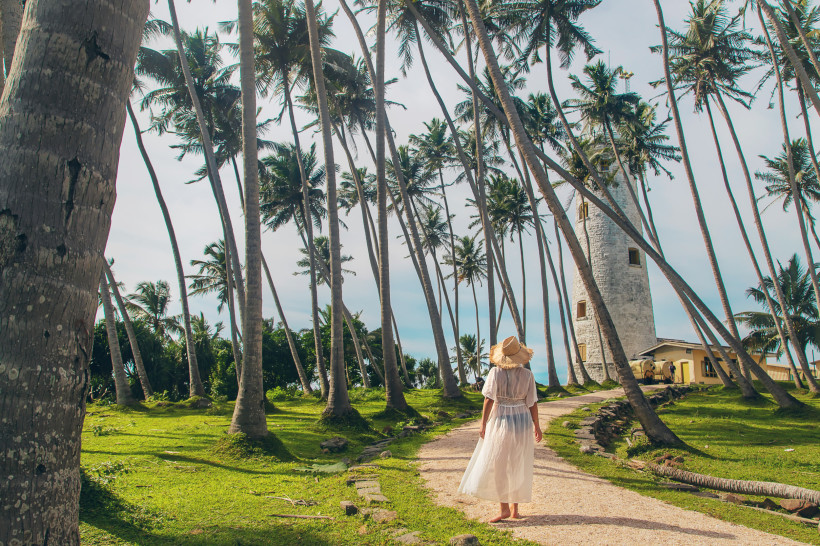 Galle, Sri Lanka Frau in weißem Kleid und Strohhut spaziert durch Palmen zum historischen Leuchtturm von Galle in Sri Lanka