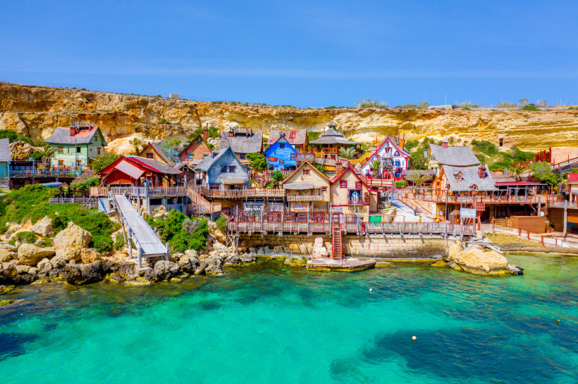 Bunte Holzhäuser des Popeye Village auf Malta an der Anchor Bay mit türkisblauem Wasser und Felsküste im Hintergrund