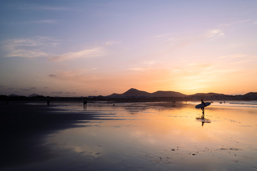Lanzarote Surfer mit Brett läuft bei Sonnenuntergang über einen spiegelnden Strand. Im Hintergrund sind Silhouetten von Bergen und Menschen im Abendlicht zu sehen.
