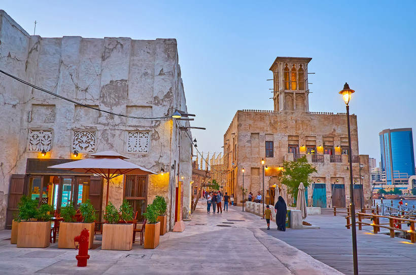 Szene in der historischen Altstadt von Dubai bei Sonnenuntergang. Traditionelle Lehmgebäude mit kunstvoll verzierten Fenstern und einem typischen Windturm säumen die Fußgängerzone. Menschen flanieren zwischen kleinen Cafés, Lichterketten und Straßenlatern