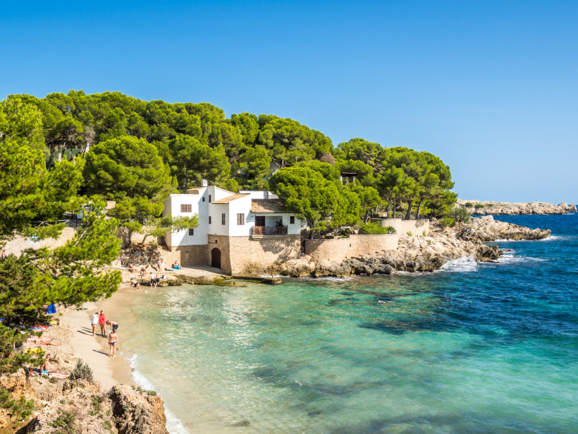 Cala Ratjada, Mallorca Kleine Badebucht mit weißem Haus und grünen Pinien bei Cala Ratjada auf Mallorca, Menschen genießen das klare Meer und den Strand