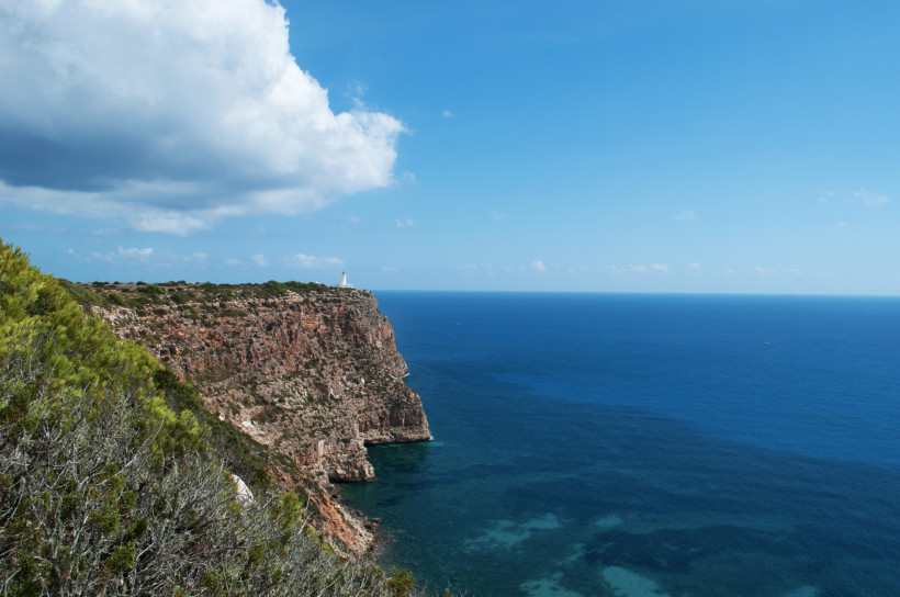 Formentera Steilküste mit rotbraunen Felsen und grüner Vegetation auf Formentera, im Hintergrund ein weißer Leuchtturm auf einer Klippe. Unten breitet sich das tiefblaue Mittelmeer aus, am Himmel ziehen weiße Wolken vorbei.