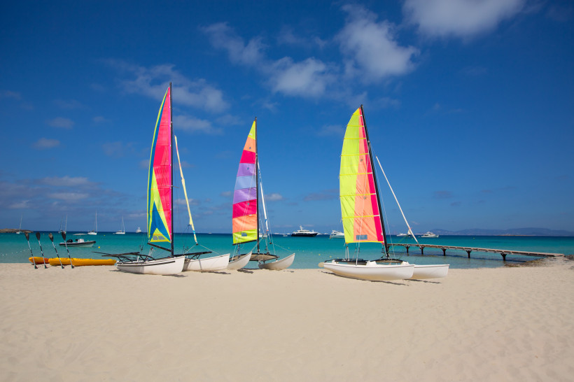 Formentera Drei kleine Segelboote mit bunten Segeln stehen am weißen Sandstrand vor türkisblauem Meer. Im Hintergrund sind Yachten und ein Holzsteg zu sehen, darüber ein blauer Himmel mit einigen kleinen Wolken.