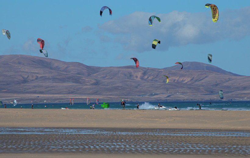 Lanzarote Weitläufiger Sandstrand mit vielen Kitesurfern und bunten Kites in der Luft. Im Hintergrund ragen braune Hügel unter blauem Himmel auf