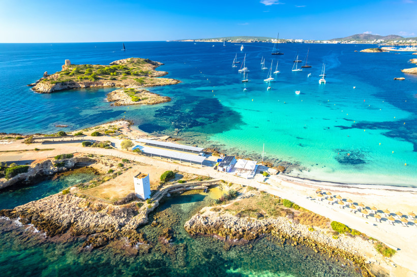 Luftaufnahme der Cala Xinxell mit türkisfarbenem Wasser, Sandstrand, felsiger Küste und mehreren Segelbooten in der Bucht