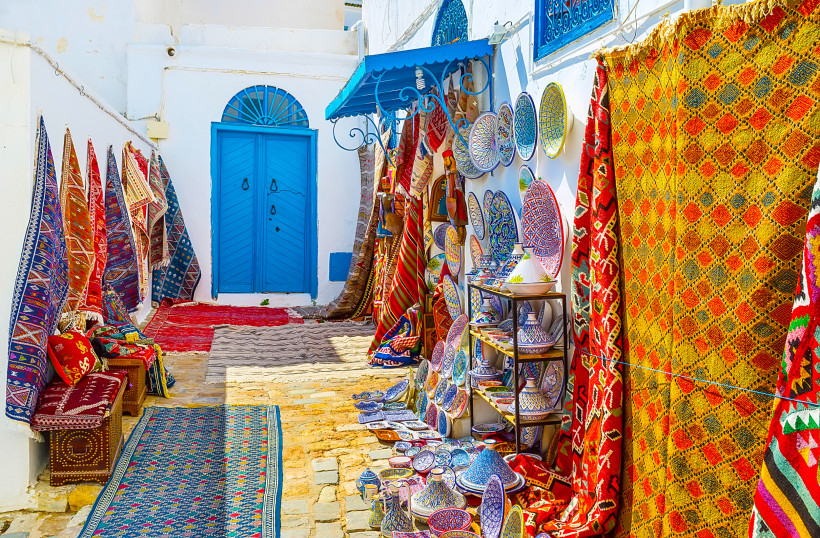 Eine enge Gasse in Sidi Bou Said, Tunesien, gesäumt von weißen Hauswänden mit einer strahlend blauen Tür. Links und rechts der Gasse hängen bunte Teppiche und Decken, die die Straße in ein Farbenmeer verwandeln. Dazwischen sind Töpferwaren und traditionel
