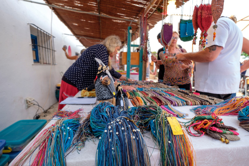 Formentera Marktstand mit einer großen Auswahl an handgefertigten, farbenfrohen Armbändern und Schmuck aus Garn. Menschen betrachten die Waren, ein Strohdach spendet Schatten