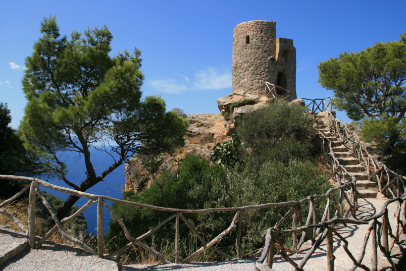 Mallorca - Torre del Verger Das Bild zeigt eine alte Wachturm-Ruine auf einer Felskuppe hoch über dem Meer. Der Turm ist rund, aus groben Natursteinen gebaut, mit kleinen Fenstern und einer hölzernen Balustrade, die sich den Felsen entlang schlängelt. Der Himmel ist strahlend blau,