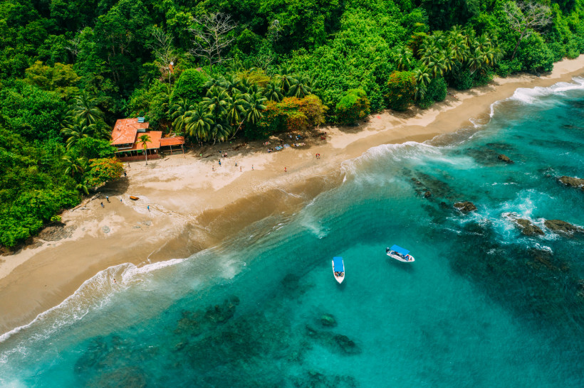 Costa Rica Luftaufnahme eines tropischen Strandes mit türkisfarbenem Wasser, zwei Booten im Meer und einem Gebäude mit rotem Dach am Rand des dichten Regenwaldes