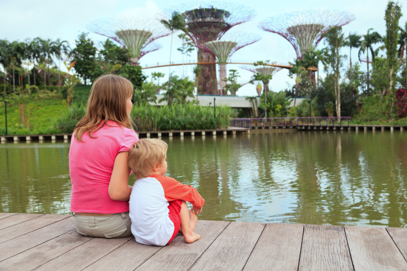Singapur Ein Mädchen und ein kleiner Junge sitzen auf einem Holzsteg an einem ruhigen See in Singapur. Im Hintergrund sind die futuristischen Supertrees von Gardens by the Bay zu sehen. Die Kinder blicken ruhig auf das Wasser und die üppige tropische Vegetation.