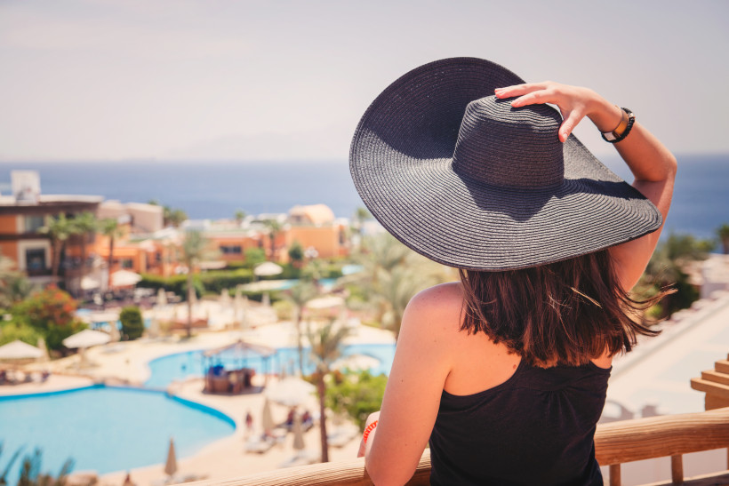 Ägypten - Sharm el Sheikh Eine Frau mit dunklem Haar und großem Sonnenhut blickt von einem Balkon auf ein weitläufiges Resort mit Pools, Palmen und orangefarbenen Gebäuden. Im Hintergrund ist das tiefblaue Meer zu sehen. Die Szene wirkt entspannt und sommerlich.