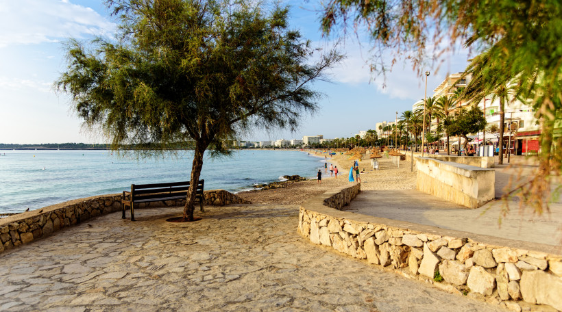 Strandpromenade auf Mallorca mit Natursteinpflaster, Sitzbank unter schattigem Baum und Blick auf das ruhige Mittelmeer entlang der Küste