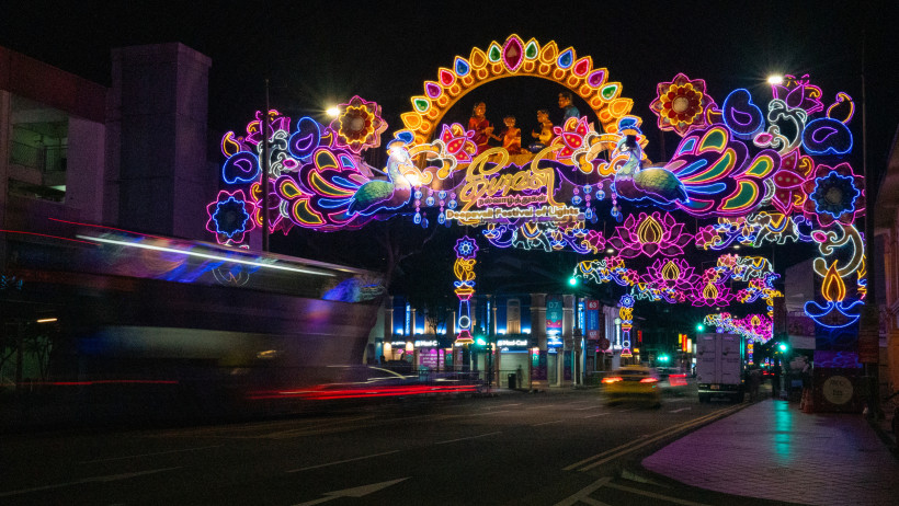 Singapur Leuchtend bunte Deepavali-Straßendekorationen in Singapur bei Nacht. Die Straße ist mit aufwendigen, farbenfrohen Lichtinstallationen überspannt, die Pfauen, Blumen und hinduistische Figuren zeigen. Im Vordergrund fahren Autos und ein Doppeldeckerbus durc